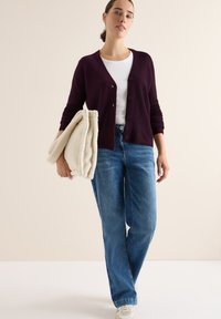 Lila gestrickte Strickjacke mit Knöpfen, weißes T-Shirt und blaue Jeans. Hält eine cremefarbene, strukturierte Decke. Lässiger Schnitt und entspannter Stil.
