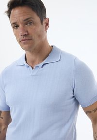 Polo de manga corta azul claro hecho de material suave. Presenta un cuello plano y puños acanalados, con una textura lisa y líneas limpias.