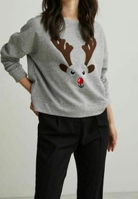 Sudadera gris con un diseño de reno con cuernos marrones, ojos negros y una nariz roja; combinada con pantalones negros.