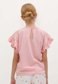 Rosa Kurzarmshirt mit Rüschensäumen und Spitzenakzenten an den Schultern. Verfügt über ein kleines Schlüsselloch am Rücken und einen Knopfverschluss.
