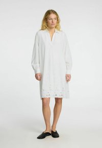 Bílé šaty s límečkem a dlouhými rukávy. Obsahují krajkový lem broderie anglaise. Modelka má na sobě černé nazouvací boty.