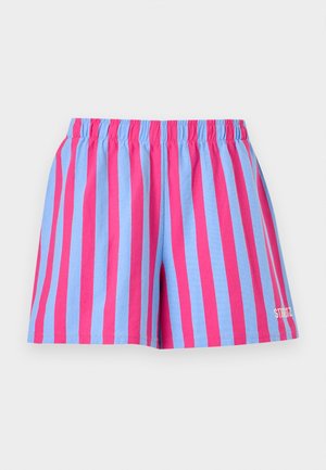 Person iført solbriller, oversize blå T-shirt med stor pink blomstergrafik, stribede pink og blå shorts, hvide sokker og røde snørebåndsstøvler.