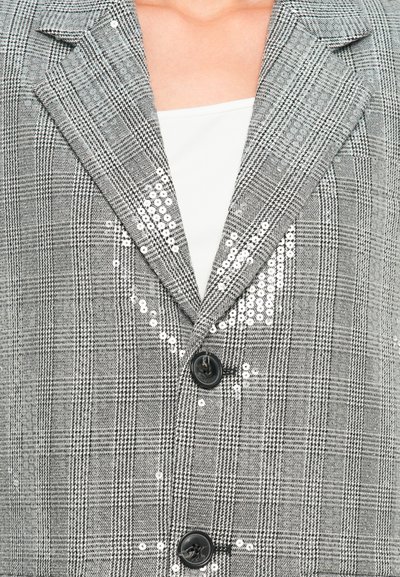 Blazer a quadri grigio con bottoni neri e sottili accenti di paillettes argento indossato sopra una maglietta bianca, con particolare attenzione all'area del petto e del colletto.