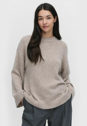 Vaaleanbeige neulepusero, jossa on pyöreä kaula-aukko ja rento malli. Ominaisuuksiin kuuluvat leveät hihat, joissa on ribbireunukset sekä helma. Yhdistetty harmaisiin housuihin.