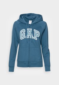 Felpa blu con zip realizzata in tessuto morbido, caratterizzata da un grande logo "GAP" in bianco, tasche a marsupio e cappuccio regolabile con cordino.