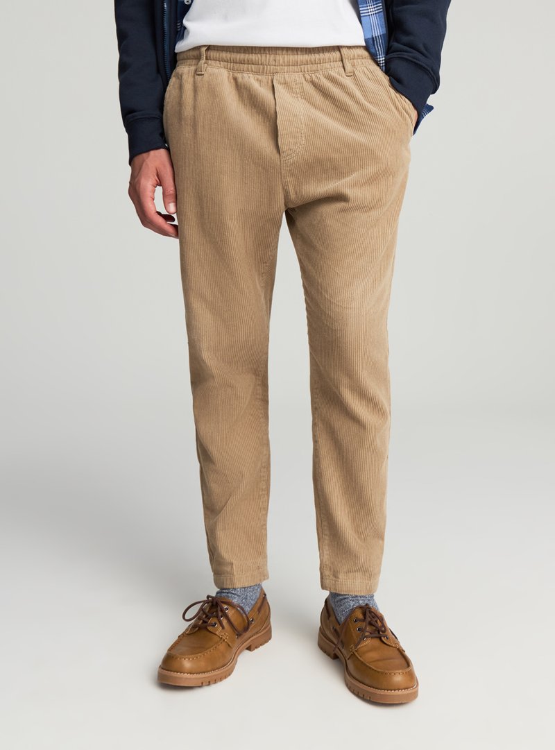 Pantaloni in velluto a coste beige con vestibilità comoda, caratterizzati da una gamba affusolata e una vita elasticizzata. Abbinati a scarpe marroni e calzini blu.