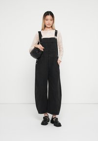 Črne denim naramnice širokih nogavic z nastavljivimi trakovi, v kombinaciji z dolgimi čipkastimi rokavi in črnimi debelimi čevlji, ki držijo majhno torbico.