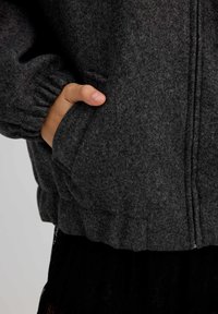 Chaqueta de forro polar gris con puños fruncidos y cremallera delantera. Mano izquierda descansando en el bolsillo lateral, mostrando su textura suave y detalles sutiles.