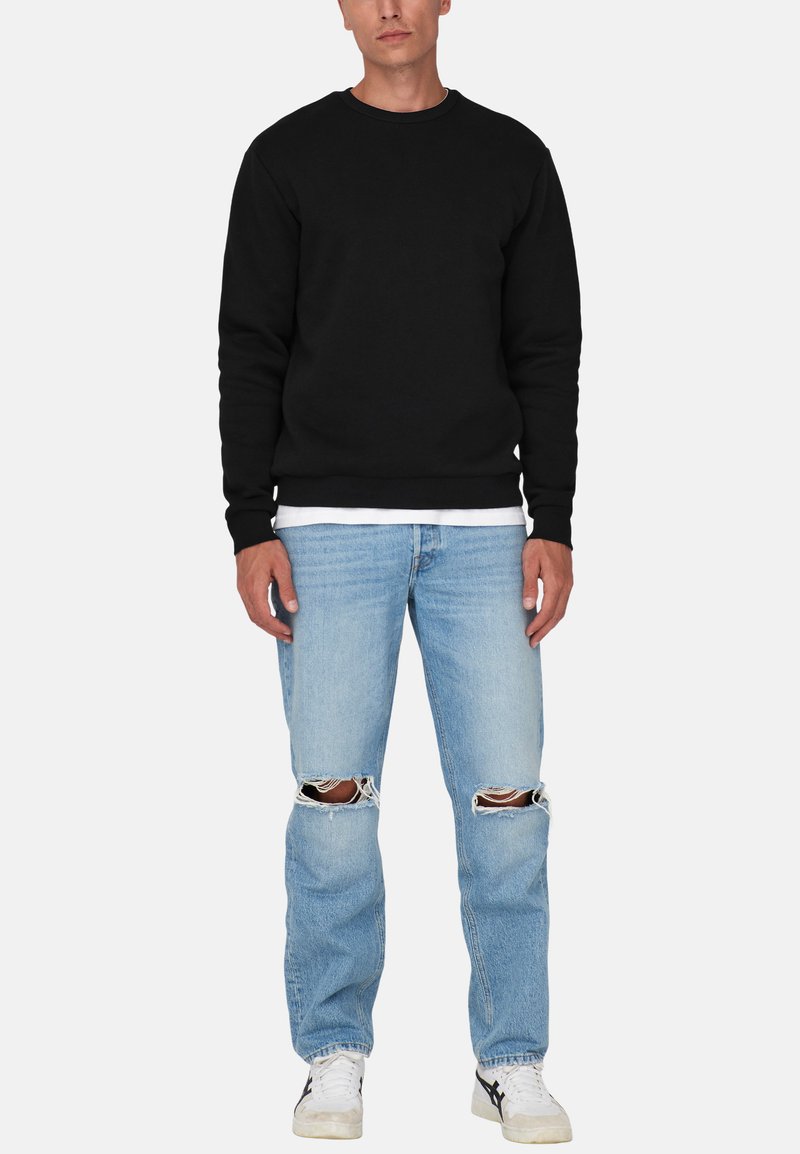 Mand står iført en sort langærmet sweatshirt, hvid undertrøje, lyseblå ripped jeans ved knæene og hvide sneakers.