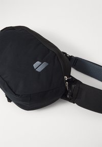 Black fabric shoulder bag with a rounded shape, featuring a zipper closure and a grey logo. Adjustable strap with textured accents.