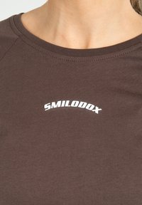 Brun tætsiddende t-shirt med rund halsudskæring og et hvidt buet logo "SMILODOX" på brystet. Blødt, strækbart tekstil med glat overflade.