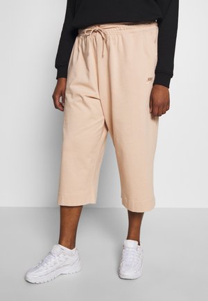 Person iført beige brede korte bukser med snøre og hvide sneakers, stående mod en ensfarvet lys baggrund.