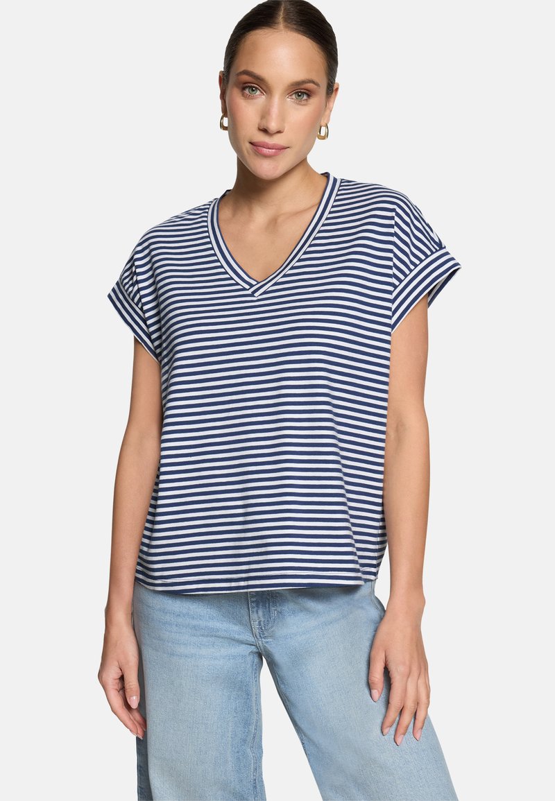 Femme portant des boucles d'oreilles créoles dorées, un t-shirt à manches courtes à col en V rayé bleu marine et blanc, et un jean bleu clair sur fond blanc.