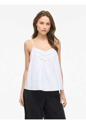 Ung kvinde med langt brunt hår iført en hvid ærmeløs top med blonder og løse sorte bukser, stående mod en ensfarvet baggrund.