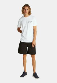 Camiseta blanca con texto gráfico negro, combinada con pantalones cortos negros y zapatillas deportivas oscuras. Ajuste casual y tejido ligero.