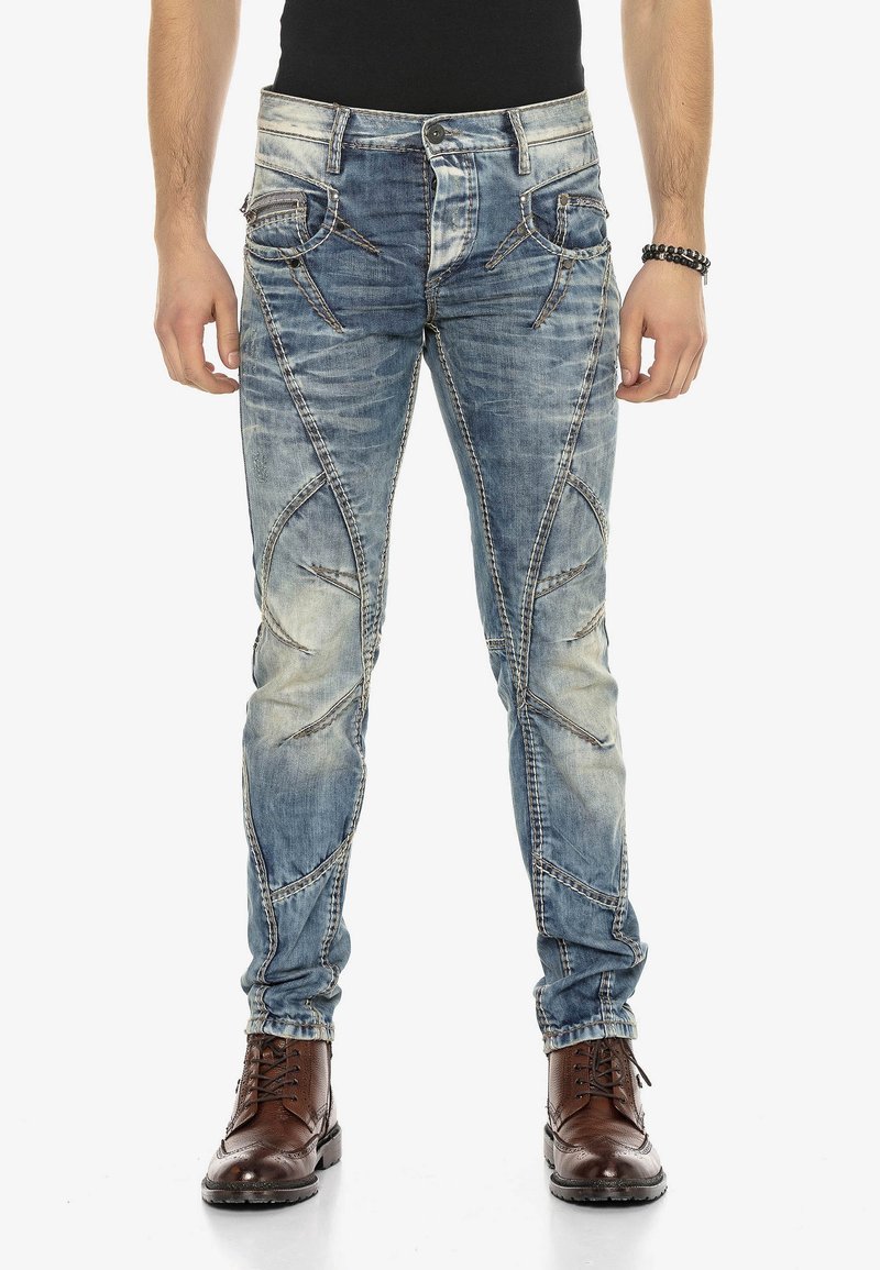 Vaqueros de denim azul desgastados con patrones de costura intrincados, cinco bolsillos y un diseño de corte ajustado. Llevados con botas marrones de cordones.