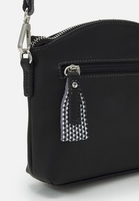 Černá kožená crossbody taška s kulatým vrškem, která má přední kapsu na zip a tkanou černo-bílou ozdobu v podobě třásní. Detail v lesklém stříbrném kovu.