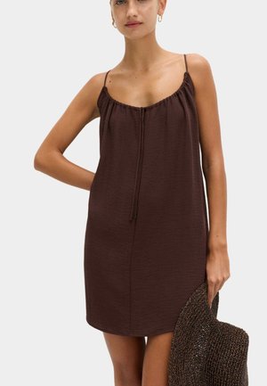 Frau trägt ein ärmelloses, braunes Minikleid mit dünnen Trägern und hält einen breitkrempigen, gewebten Hut in ihrer rechten Hand.