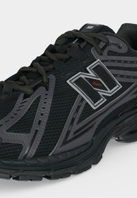 Μαύρο και γκρι αθλητικό παπούτσι New Balance με διαπνέον δίχτυ, συνθετικά επικάλυψη και το λογότυπο με το γράμμα "N" στη πλευρά κοντά στα κορδόνια.