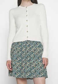 Biały prążkowany kardigan z długimi rękawami, zdobiony rzędem złotych guzików z przodu, zestawiony z wielokolorową spódnicą w wzory.