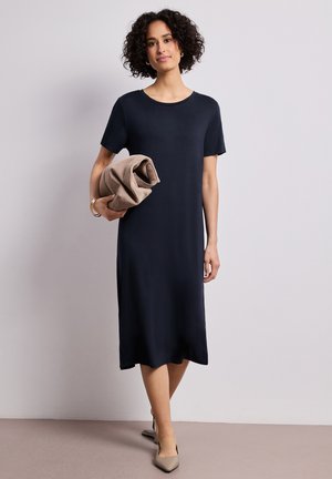 Kvinde med krøllet hår iført en navyblå midi-kjole og beige hæle, der holder en foldet beige frakke mod en ensfarvet baggrund.