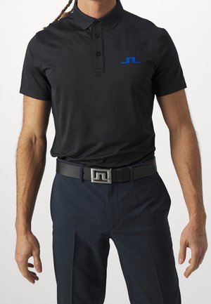 Mann trägt ein schwarzes kurzärmeliges Poloshirt mit blauem Logo, marineblaue Hose und schwarzen Gürtel mit silberner Schnalle, steht vor einem einfarbigen Hintergrund.