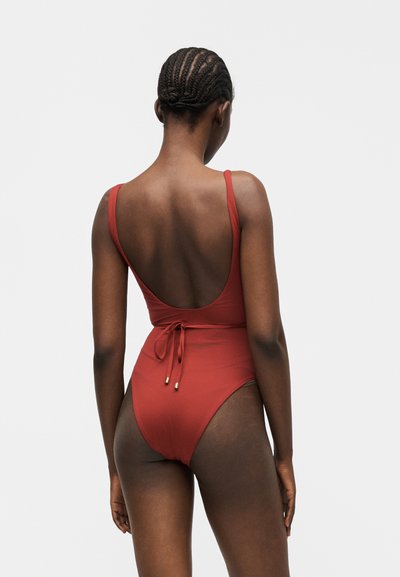 Γυναίκα με πλεξούδες που φοράει ένα ολόσωμο μαγιό σε σκουρόχρωμο κόκκινο με χαμηλή πλάτη και δεμένη ζώνη, στέκεται μπροστά σε ένα απλό ανοιχτό γκρι φόντο.