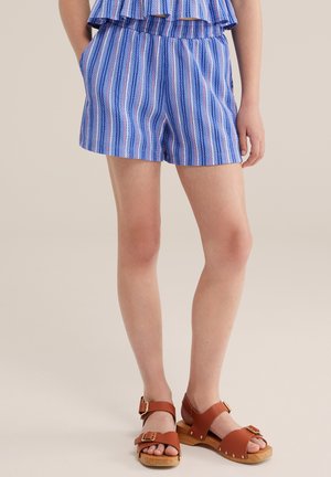 Žena oblečená v modro-biele pruhované šortky a hnedé sandále s drevenou podrážkou a s prackami, stojaca na jednoduchom pozadí.