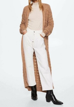 Femme portant un col roulé beige, un long cardigan marron, un pantalon blanc taille haute avec des boutons, et des bottines noires à talons, les mains dans les poches.