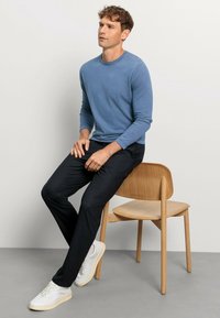 Hellblaues Langarmhemd kombiniert mit dunklen Hosen, sitzt auf einem Holztstuhl. Trägt weiße Sneakers und hat einen neutralen Hintergrund.