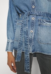 Denim jakna v svetlo modri barvi z pasom, gumbom za zapiranje in žepom na prsih. Ima rahlo obrabljen videz.