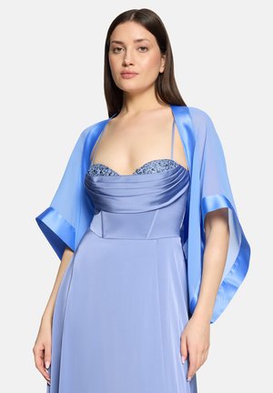 Frau mit langen dunklen Haaren, die ein hellblaues Kleid aus Satin und Chiffon trägt, mit einem transparenten Schal, der über die Schultern drapiert ist, stehend vor weißem Hintergrund.