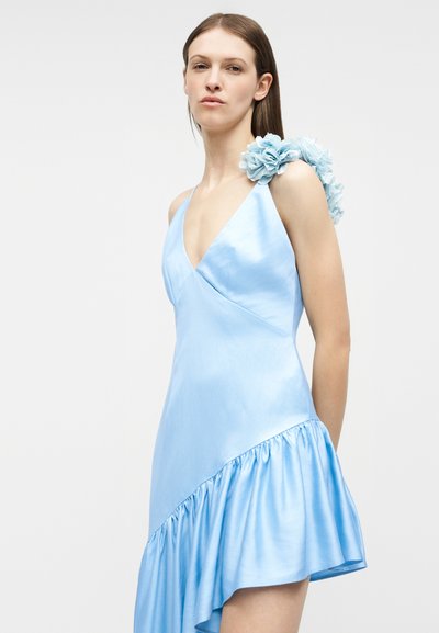 Γυναίκα που φοράει ανοιχτό μπλε σατέν φόρεμα με ασύμμετρο βολάν στο τελείωμα και τρισδιάστατες ανθικές λεπτομέρειες στον έναν ώμο.