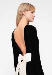 Vestido de terciopelo negro con una espalda abierta profunda y mangas largas. Presenta un gran lazo de satén crema en la cintura. Textura suave y diseño ajustado.