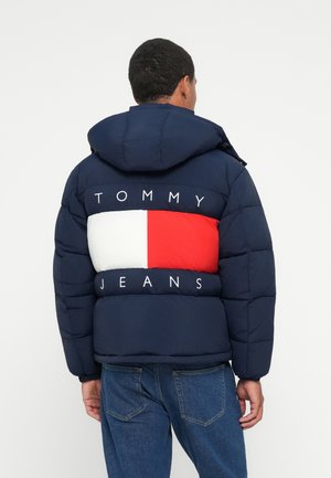 Doudoune navy avec un grand motif en blocs rouges et blancs, arborant le logo "TOMMY JEANS" en blanc dans le dos et une capuche.