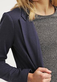 Blazer azul marino con mangas largas y corte entallado, superpuesto sobre una blusa texturizada gris claro, destacando las líneas limpias y el diseño moderno.