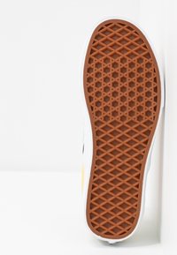 Semelle en caoutchouc d'une basket blanche avec un motif en forme de gaufre marron, photographiée sur fond blanc.