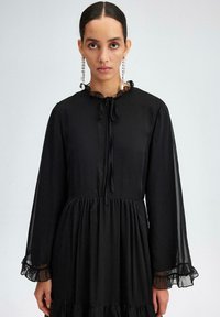Black dress with long, sheer sleeves, ruffled neckline, and tie closure. Flared skirt with gathered detailing at the waist.