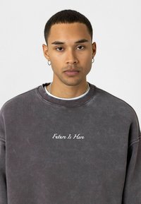 Grijze sweatshirt van zacht materiaal, met een ronde hals en raglanmouwen. "De Toekomst Is Hier" is in het wit geborduurd op de borst.