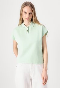 Muntgroene polo met korte mouwen en een kraag, met gouden knopen en een gestructureerde stof. Gecombineerd met witte broek.