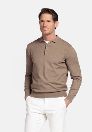 Man draagt een bruine lange mouw polo trui en witte broek, staat met handen in de zakken en kijkt lichtjes opzij.