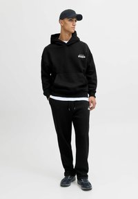 Schwarzer Hoodie und Jogginghose aus weicher Stoffqualität, mit einer Fronttasche und Logodetails. Abgerundet durch eine schwarze Mütze und sportliche Schuhe.