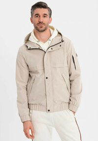 Mann mit kurzem dunklem Haar trägt eine beige Kapuzenjacke über einem weißen Hoodie und weißen Hosen und steht vor einem schlichten weißen Hintergrund.