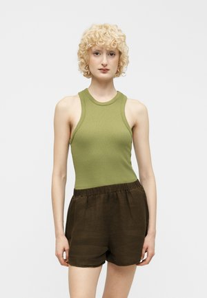 Persona de cabello rizado y rubio, vistiendo una camiseta sin mangas verde oliva y pantalones cortos sueltos de color marrón oscuro, de pie frente a un fondo liso y claro.