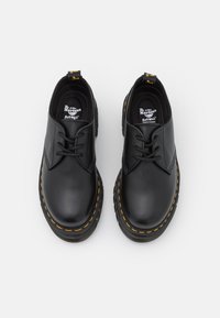 Chaussures Dr. Martens en cuir noir avec des coutures jaunes et des lacets noirs, vues de dessus sur un fond blanc.