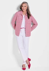 Rosa und weiße Hahnentrittmuster-Jacke, weißes Oberteil, weiße Hose und fuchsiafarbene Absätze. Die Jacke hat einen kragenförmigen Ausschnitt und Knöpfe.