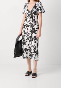 Robe portefeuille florale noir et blanc avec des manches courtes à volants, un décolleté en V et un nœud à la taille. Associée à des sandales glissantes noires et un sac.