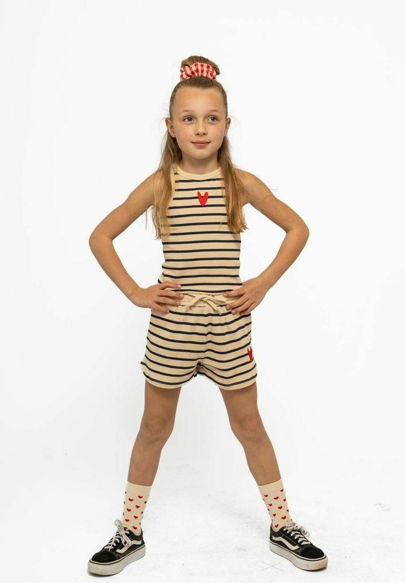 Combinaison rayée beige et marine avec un détail en forme de cœur rouge ; baskets noires et chaussettes crème avec des cœurs rouges ; cheveux retenus par un chouchou à carreaux rouges.