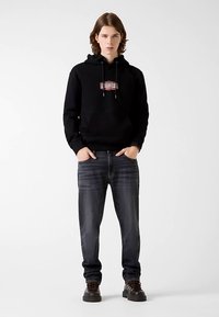 Sweat à capuche noir avec cordons de serrage ajustables et logo sur le devant, accompagné d'un jean en denim foncé et de chaussures noires à lacets avec semelles texturées.