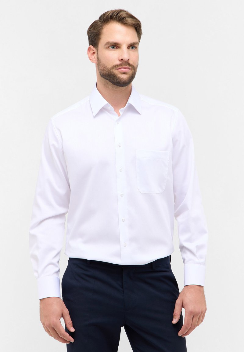 Camicia bianca a maniche lunghe con colletto, taschino sul petto, e texture liscia, abbinata a pantaloni blu scuro.